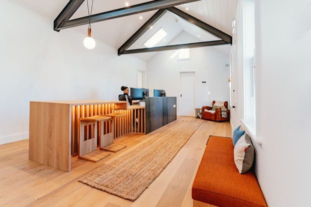 Bright contemporary open-plan interior featuring a timber reception or work counter with integrated vertical slat detailing and warm concealed lighting, multiple computer workstations, light timber flooring, a large natural-fibre rug, and built-in orange window seating. The room has a high pitched ceiling with exposed dark structural beams, skylights, white wall panelling, and generous glazing. A small lounge area with leather armchairs is visible at the rear.