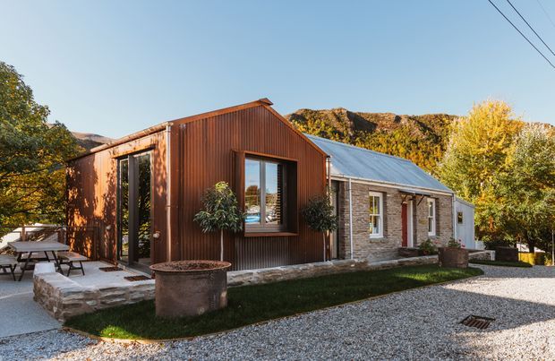 Exterior view of a renovated single-storey building combining a corrugated metal-clad gabled extension with an older stone masonry cottage. The home sits beside a gravel driveway and landscaped outdoor area, with surrounding trees and rocky hills visible in the background. Large glazed doors and windows connect the extension to a small paved terrace.