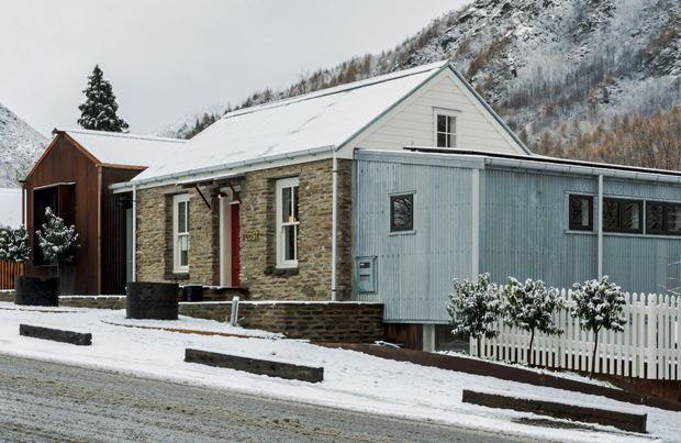 A small single-storey cottage in a snowy alpine setting. The main dwelling has a steep gabled roof, pale stone masonry walls with contrasting white trim, and a red entry door. A corrugated grey metal-clad extension projects across the front, with narrow windows and a raised deck enclosed by a white picket fence. Snow-covered mountains and trees form the background.