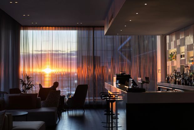 A moody, sophisticated interior combining a lounge and home bar. Floor-to-ceiling sheer curtains frame a dramatic sunset, while dark flooring, charcoal walls and ceiling, a compact bar counter with stools, illuminated bottle shelving, and comfortable lounge seating create an intimate entertaining space.