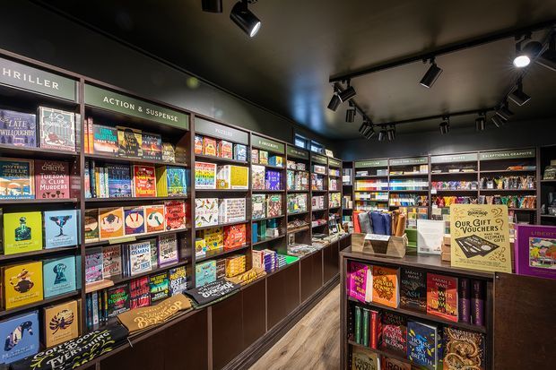 Interior view of a dark, stylish bookstore with extensive floor-to-ceiling shelving filled with books and magazines. Track lighting illuminates the displays, while dark cabinetry, black ceilings, and timber-look flooring create a dramatic retail environment. A central display island and genre signage are visible.