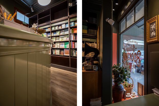 A richly detailed, dark-toned retail interior viewed from behind a paneled service counter. Built-in shelving displays numerous packaged products, while a glazed storefront door or window reveals signage outside. The space features dark walls and joinery, warm timber flooring, a framed artwork, pendant and recessed lighting, and a potted plant near the entrance. The image appears to show a boutique shop or café-style commercial space.
