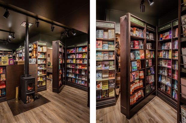 A compact, dark-toned retail bookstore or magazine shop interior with floor-to-ceiling shelving densely stocked with books and magazines. The space features warm wood-look flooring, dark walls and ceiling, black track lighting, tall dark timber shelving, and a freestanding wood-burning stove with logs in the foreground.