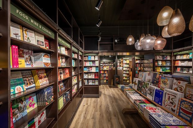 A warmly lit specialty bookstore or magazine shop interior, with dark timber shelving and built-in display joinery lining the walls and central aisles. Shelves are densely stocked with books, magazines, and printed publications, while woven pendant lights hang from a dark ceiling. The space has a polished timber-look floor and an intimate, curated retail atmosphere.