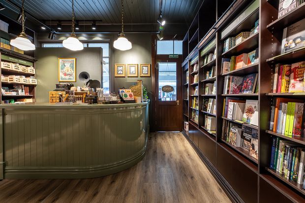 A warmly lit, compact bookshop interior with a curved dark green counter, extensive dark timber bookshelves filled with books, pendant lights, framed artwork, and a central glazed entry door. The space has timber-look flooring, dark painted timber ceiling boards, and a cosy retail atmosphere.
