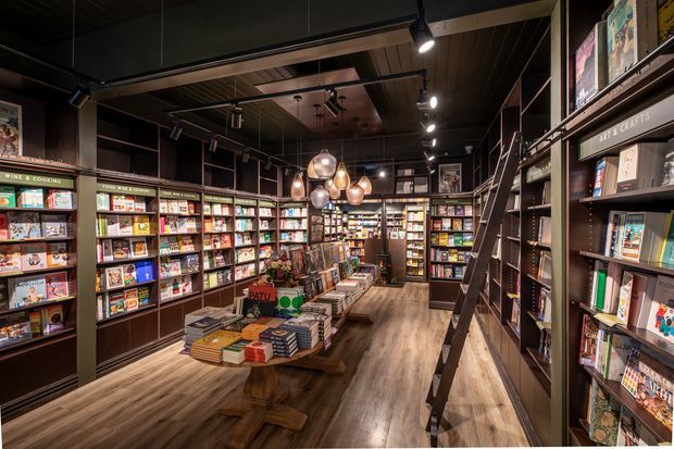 A warm, dark-toned independent bookstore or commercial retail space with floor-to-ceiling shelving filled with books, central display tables, timber-look flooring, a dark ceiling, track lighting, and clustered pendant lights. A rolling ladder provides access to the tall shelving along the right wall.