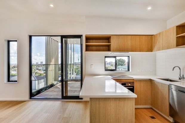 Bright contemporary kitchen with warm timber cabinetry, white benchtops and splashback, a central island, stainless-steel appliances, and recessed ceiling lighting. Full-height sliding glass doors open to a narrow balcony or terrace with urban views, bringing in abundant natural light.