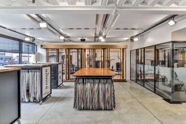 Bright contemporary commercial showroom with an open-plan layout, polished concrete flooring, exposed white ceiling structure, track lighting, glass-partitioned display rooms, and fabric sample displays arranged around a central timber-topped island.