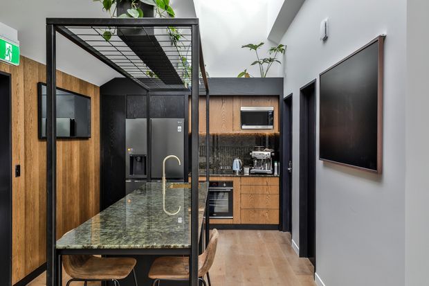 Contemporary compact kitchen and dining space with warm timber wall panelling and cabinetry, dark charcoal cabinetry and appliances, a green-toned stone island with integrated seating, and a black metal-framed overhead plant structure. A wall-mounted television is visible on the right, and a large skylight or clerestory opening brings in natural light. The space has a refined, high-contrast palette with indoor planting.