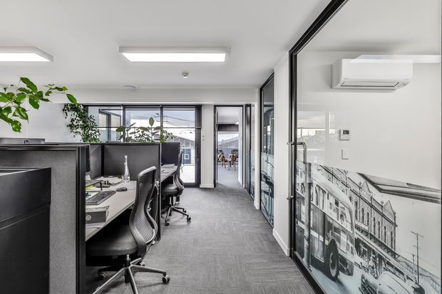 Contemporary open-plan commercial office interior with rows of workstations, black-framed glazed partitions, grey carpet flooring, white ceilings, linear fluorescent lighting, potted greenery, and a large monochrome architectural mural. A compact meeting or circulation area is visible beyond the workstations.