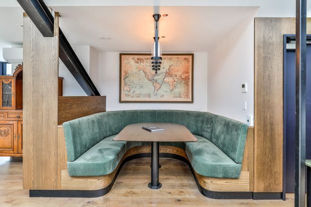 Interior view of a built-in curved banquette dining nook with teal upholstered seating, a central pedestal table, and timber-clad structural partitions. The space combines warm wood flooring and joinery with a black steel diagonal beam and a distinctive pendant light. A framed world map and an adjacent vintage timber cabinet add character to the open-plan room.