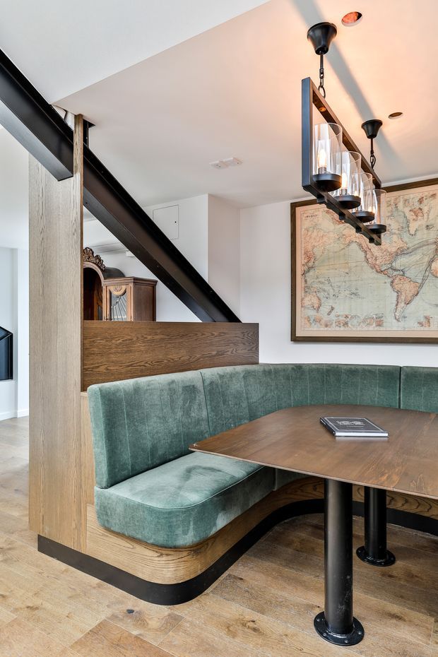 A refined dining nook with a curved, upholstered banquette in deep green velvet surrounding a dark timber table. The space features light timber flooring and wall panelling, black metal structural beams and table supports, a linear black pendant light with glass cylinders, and a large framed vintage-style world map. An ornate timber cabinet is partially visible behind the divider, adding traditional character to the otherwise contemporary fit-out.