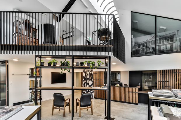 A contemporary double-height open-plan living and dining area with a mezzanine level enclosed by black vertical metal balustrades. The space features a black-framed open shelving partition with plants, a small dining setting, timber slatted wall detailing, dark kitchen cabinetry, polished concrete flooring, and large black-framed architectural artwork. A curved glazed roof or skylight structure brings natural light into the interior.