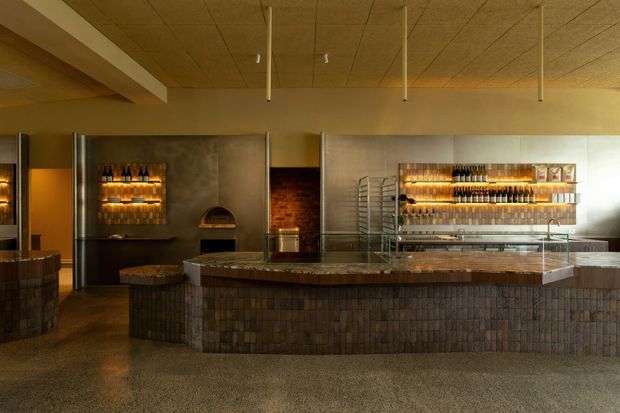 Interior view of a contemporary commercial bar or hospitality venue, featuring a long curved bar counter clad in dark textured tile or brick, a stone-look benchtop, illuminated wall-mounted bottle shelving, exposed brick accents, and a warm timber-toned ceiling with suspended linear elements. The subdued amber lighting creates a refined, moody atmosphere.