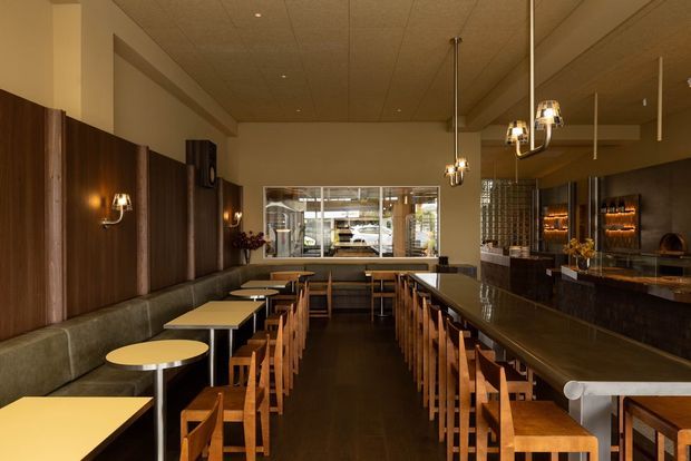 A warmly lit contemporary restaurant or bar interior with a long central counter lined with wooden stools, small light-coloured dining tables, and built-in upholstered banquette seating along a dark timber-paneled wall. Pendant lights hang above the counter and tables, while a back bar with illuminated shelving and bottles is visible at the right. A large internal window opens views toward a service or kitchen area at the rear.