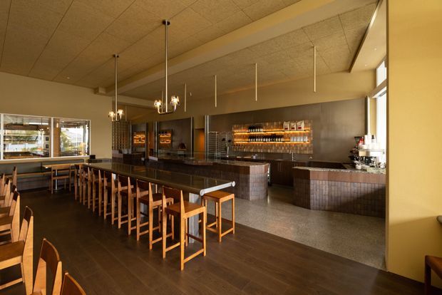 Warmly lit contemporary commercial bar and dining space with a long dark counter, timber bar stools and chairs, tiled service counters, illuminated bottle shelving, stainless-steel appliances, and a textured acoustic ceiling with suspended pendant lights. Large glazing opens the venue to the exterior.