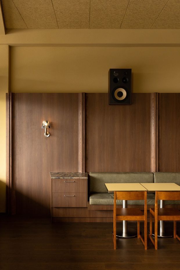 A warmly lit, richly detailed interior featuring dark timber wall panelling with vertical battens, integrated olive upholstered banquette seating, a small wooden dining table with matching chairs, a wall-mounted speaker, and a decorative wall sconce. The space has dark timber flooring and a textured acoustic-panel ceiling, creating an intimate, refined atmosphere with mid-century-inspired detailing.