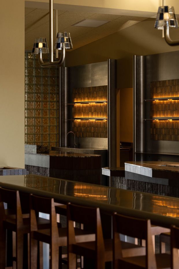 A moody, refined hospitality-style interior featuring a long dark dining or bar counter with timber chairs in the foreground. Behind it are dark metallic wall panels, warm illuminated textured tile feature walls, a glass-block partition, built-in counters with sinks, and sculptural chrome-and-glass pendant lights. The palette is predominantly deep brown, bronze, black, and warm amber.