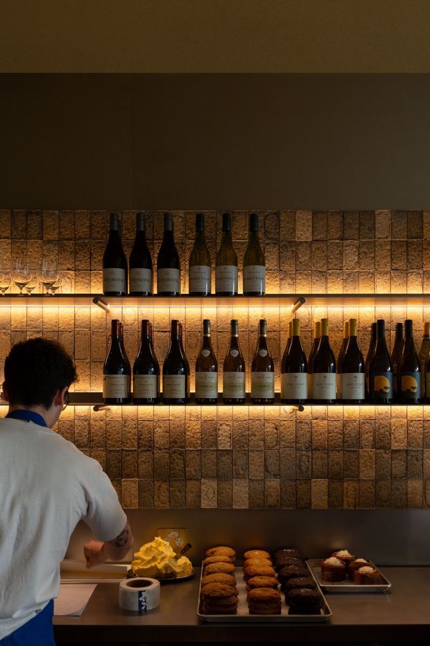 A warmly lit hospitality space featuring wall-mounted shelves displaying wine bottles and glasses against a textured brown tiled backsplash. A counter below holds coffee and baked goods, with a person partially visible in the foreground. Integrated shelf lighting creates a moody, amber-toned atmosphere.
