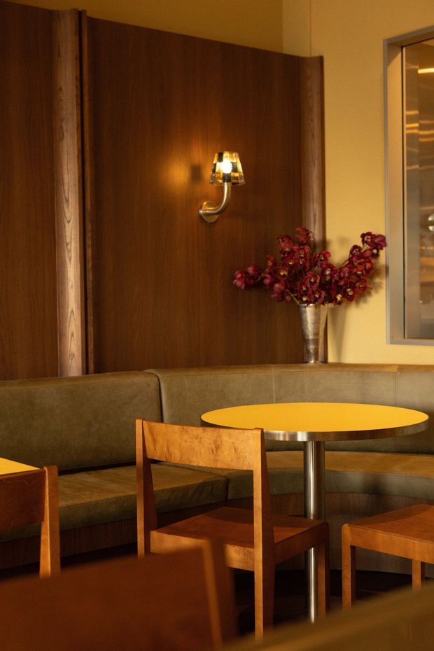 A warmly lit hospitality-style seating area with a curved upholstered banquette, wood dining chairs, and a round yellow tabletop. Dark timber wall panelling, a wall sconce, a vase of dried flowers, and a framed interior opening create a richly coloured, intimate setting.
