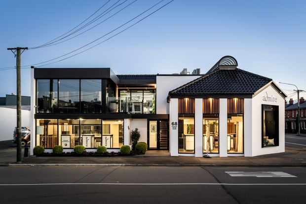 Street-facing view of a contemporary commercial building, combining a black-framed glazed two-storey wing with a white rendered, tiled-roof frontage. Large display windows reveal a warmly lit retail or showroom interior, while vertical timber-toned screening and integrated signage define the facade. Overhead utility wires, roadway markings and neighbouring urban buildings reinforce the streetscape setting.