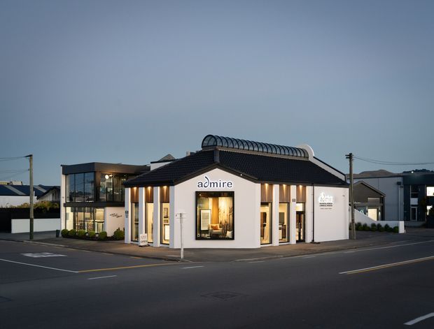 Twilight exterior photograph of a contemporary low-rise commercial building on a street corner. The building has a dark tiled pitched roof, white rendered walls, extensive glazing, illuminated display windows, and a distinctive curved rooftop skylight or canopy. Nearby modern residential or commercial buildings and utility poles are visible in the background.