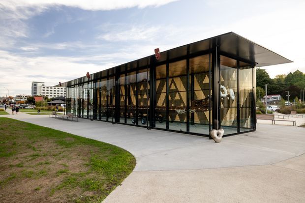A modern single-storey pavilion-like building in an open urban landscaped setting. The structure has extensive floor-to-ceiling glazing within a black metal frame, a broad flat overhanging roof, and patterned opaque interior screens or panels visible through the glass. Concrete paths and paved areas surround the building, with lawns, benches, trees, and distant commercial buildings in the background.