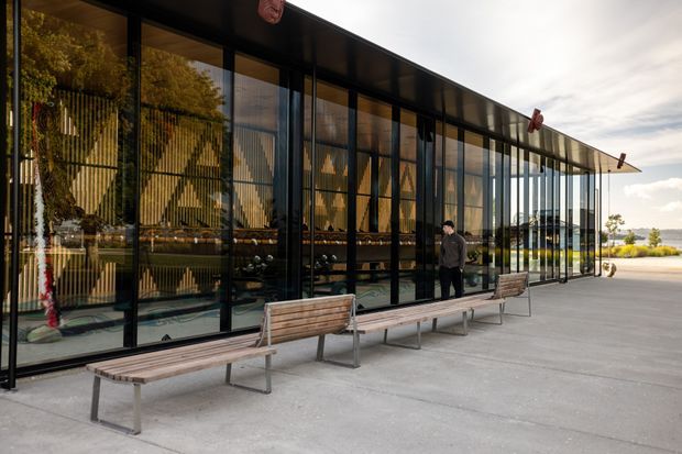 Exterior view of a long, low contemporary pavilion with extensive floor-to-ceiling dark-framed glazing. The glass reflects surrounding trees and reveals a warm timber-lined interior, while a broad concrete forecourt with timber-and-metal benches runs along the facade. A landscaped waterfront or open landscape is visible in the distance.