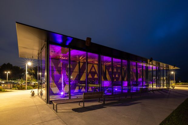Night-time exterior view of a long, low contemporary pavilion or commercial building with extensive floor-to-ceiling glazing, a broad projecting canopy, and dark structural framing. Strong purple and blue interior lighting illuminates patterned screens or graphics visible through the glass. The building sits beside a paved pedestrian forecourt with benches and exterior lighting.