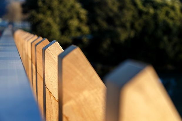Close-up view along a timber balustrade or privacy screen, with repeated vertical wooden panels and dark metal or painted supports beside a walkway or deck. The shallow depth of field emphasizes the warm wood grain and geometric rhythm, with blurred greenery in the background.