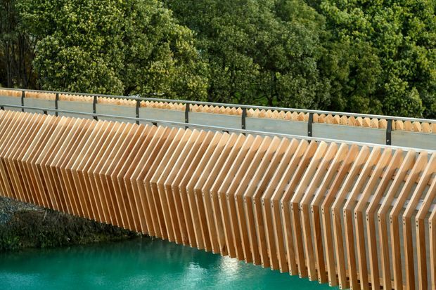 Close-up exterior view of a distinctive pedestrian bridge or bridge-like structure spanning turquoise water. Repeated warm timber ribs form a sculptural side screen or balustrade, with a grey metal upper railing and dense green trees in the background.