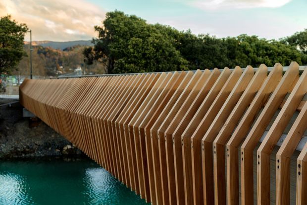 A close view of a distinctive timber pedestrian bridge or elevated walkway with repeated angled slats forming a sculptural balustrade and side structure. The bridge curves over turquoise water, with trees and hills visible in the background.