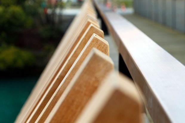 Close-up architectural detail of a timber balustrade or privacy screen beside a pale timber handrail, with shallow depth of field. The repeated angled timber elements create a rhythmic geometric pattern, while blurred greenery and blue water appear in the background.