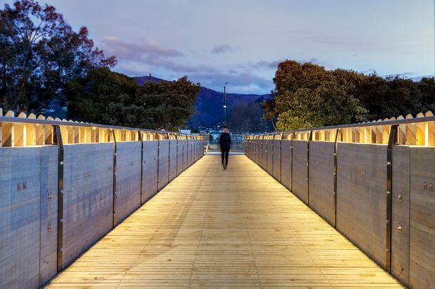 A long illuminated pedestrian bridge or elevated walkway at dusk, with warm timber decking, translucent side panels, and angular illuminated railings. A person walks toward a distant mountainous, tree-lined setting.