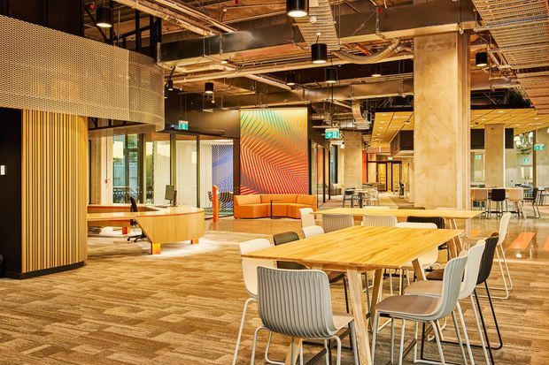 A spacious contemporary commercial interior with an open-plan dining, collaboration and lounge area. The space features exposed building services and ductwork, suspended black pendant lights, concrete columns, timber slatted screening, perforated metal wall panels, glazed meeting-room partitions, patterned carpet tiles, timber tables, and colourful lounge seating. A large geometric artwork provides a vivid focal point.