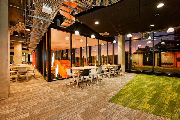 Open-plan contemporary commercial interior with exposed services, black ceiling panels, extensive black-framed glass partitions, pendant lighting, collaborative dining-style tables, and a mix of neutral and vivid green carpet tiles. Warm orange-lit meeting rooms and reflected office spaces are visible through the glazing.