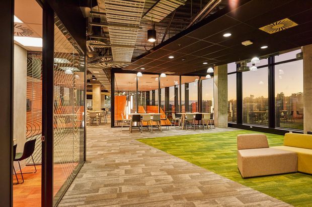 A contemporary open-plan commercial interior, likely an office or collaborative workplace, with exposed services and a patterned acoustic ceiling, black ceiling panels, extensive floor-to-ceiling glazing, meeting and dining tables, upholstered lounge seating, patterned carpet, and a bright green carpeted zone. Decorative glazed partitions and warm orange feature surfaces add visual interest, while the windows overlook an urban landscape.
