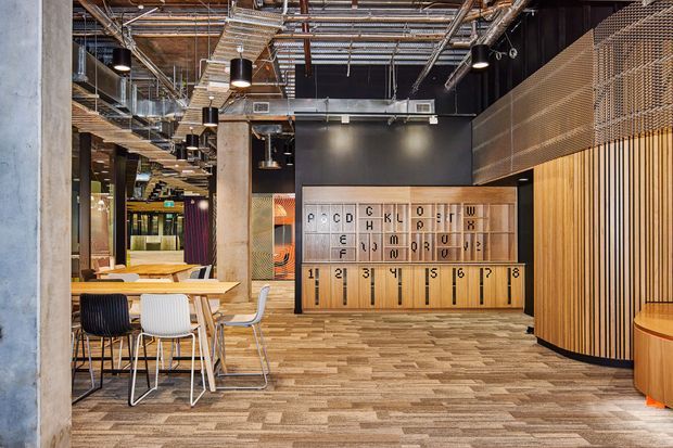 An open-plan contemporary commercial interior with exposed services, concrete columns, black ceilings, suspended cylindrical pendant lights, timber acoustic screening, and a large built-in timber feature wall with letter and number compartments. A communal dining or work table with mixed chairs sits to the left, while patterned carpet tiles cover the floor.