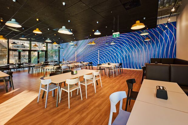 A spacious contemporary commercial dining and café interior with extensive timber-look flooring, a black ceiling, colorful pendant lights, and rows of light tables and chairs. The standout feature is a vivid blue feature wall covered in sweeping red and white curved line graphics. Glazed frontage opens toward an urban exterior, while dark upholstered booth seating occupies the right side.