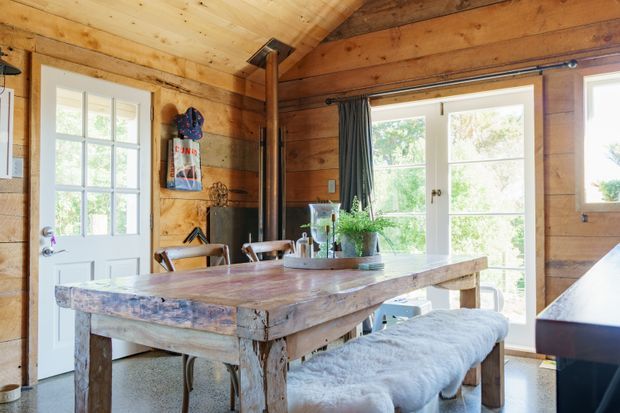 Warm rustic timber-lined dining area in a compact cabin-like interior. A substantial reclaimed timber dining table with a white upholstered bench sits beside white-framed glazed doors and windows, with a black wood-burning stove and exposed timber ceiling and beams adding character.