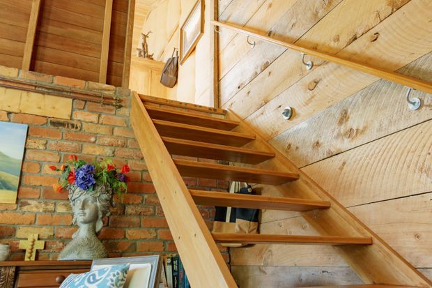 Warm rustic interior view of an open timber staircase rising beside a reclaimed brick feature wall. The stair treads, handrail, wall cladding and ceiling are natural timber, with exposed construction details and eclectic decorative elements visible below and around the stair.