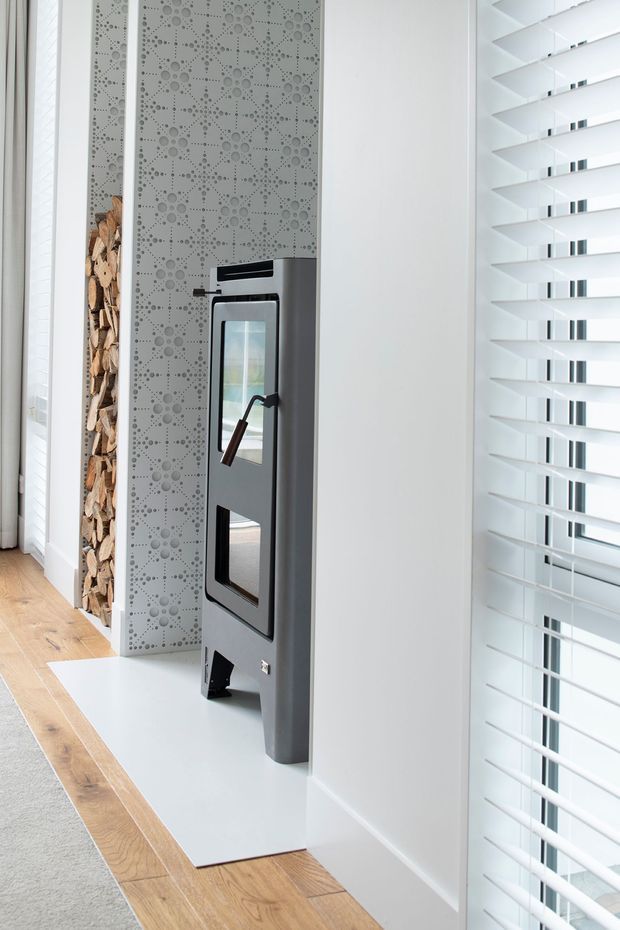 Bright contemporary interior corner featuring a freestanding dark metal wood-burning stove with a glass-fronted door, set against a decorative perforated grey feature panel. A tall recessed stack of firewood sits beside it. White walls, plantation shutters, pale timber flooring, and a light hearth create a clean, refined setting.