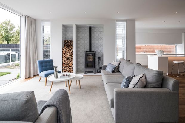 Bright contemporary open-plan living room with a large grey sectional sofa, matching armchairs, round white coffee tables, and a freestanding wood-burning stove set into a patterned tiled fireplace surround with stacked firewood niches. Floor-to-ceiling glazing opens to an outdoor area, while the adjoining white kitchen includes a peninsula with stools and timber flooring.