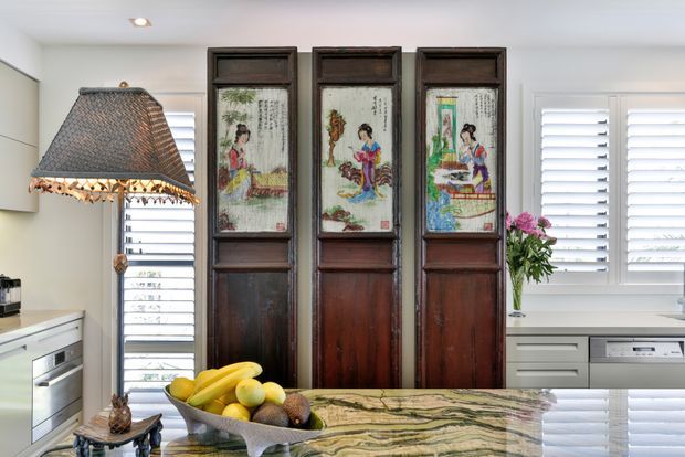 Bright, eclectic kitchen interior with white cabinetry, a glossy veined stone island benchtop, plantation shutters, and three tall dark timber decorative panels featuring painted artwork. A large woven pendant light hangs over the island, with fruit and flowers adding colour.