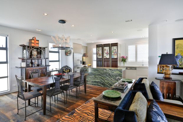 Bright open-plan dining and living area with a traditional eclectic interior. The room features a dark timber dining table and chairs, ornate display shelving, a large veined stone island or console, timber flooring, white cabinetry, a navy sofa, patterned rug, decorative lighting, and framed artwork.