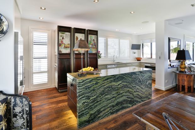 Bright, spacious kitchen with a large green veined stone island, white cabinetry, plantation shutters, and warm timber flooring. The room includes recessed ceiling lights, a decorative folding screen, and views into an adjacent living area.