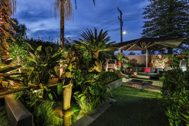 A richly planted landscaped outdoor courtyard or garden at dusk, featuring dense tropical foliage, illuminated water or sculptural features, dark paving and artificial turf, plus a covered outdoor lounge with a large umbrella, sofa seating and red cushions. The setting has a dramatic, intimate evening atmosphere.