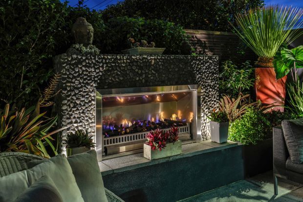 A lush, moody outdoor lounge area centred on a built-in linear fireplace. The fireplace surround is clad in textured dark pebble-like stone, with a reflective metal firebox and flame-effect lighting. Tropical planting, potted greenery, a Buddha statue, outdoor seating and a raised dark-rendered hearth create a secluded garden entertaining setting.