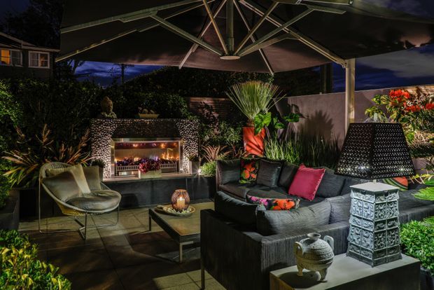 A luxurious, atmospheric outdoor lounge and entertaining area at night, arranged beneath a large cantilevered umbrella. The space features a dark sectional sofa with colourful cushions, sculptural lounge chairs, a coffee table, layered tropical planting, decorative lighting, and a built-in stone-clad outdoor fireplace with shelving and a bar-like display. The composition has a warm, dramatic resort-style ambience.