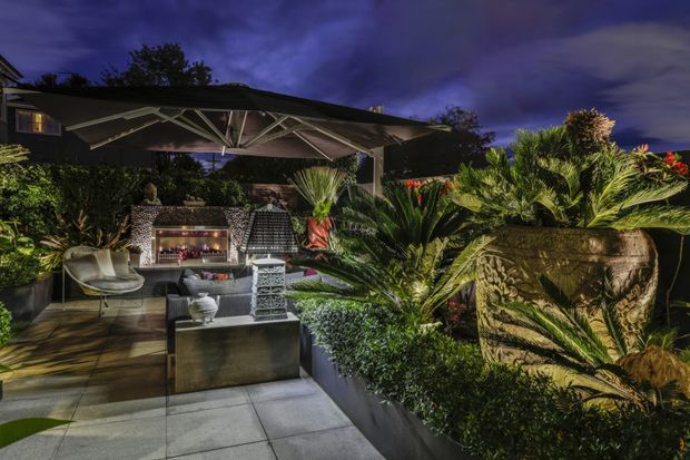 A richly landscaped outdoor entertaining area at night, featuring a large covered patio with an expansive cantilevered shade structure, outdoor lounge seating, a built-in fireplace or barbecue feature, tiled paving, dense tropical planting, and sculptural raised planters. Dramatic architectural lighting highlights the foliage and outdoor furnishings.